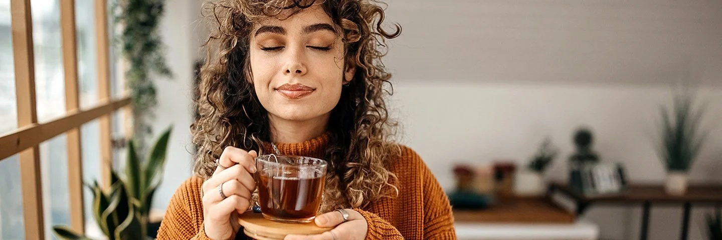 Eine Frau in einem braunen Pullover hält mit geschlossen Augen eine Tasse Tee in der Hand.