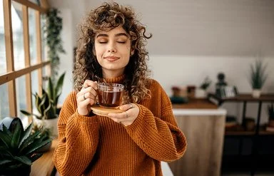 Eine Frau in einem braunen Pullover hält mit geschlossen Augen eine Tasse Tee in der Hand.