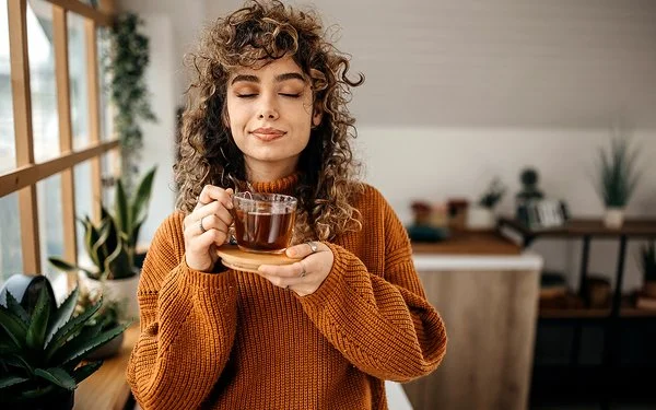 Eine Frau in einem braunen Pullover hält mit geschlossen Augen eine Tasse Tee in der Hand.