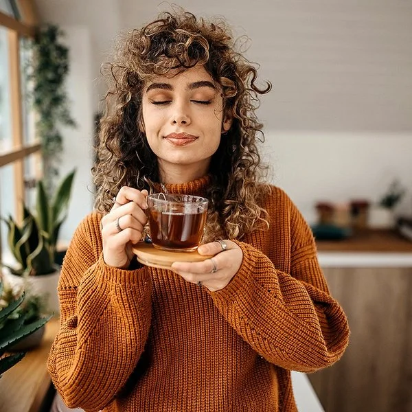 Eine Frau in einem braunen Pullover hält mit geschlossen Augen eine Tasse Tee in der Hand.
