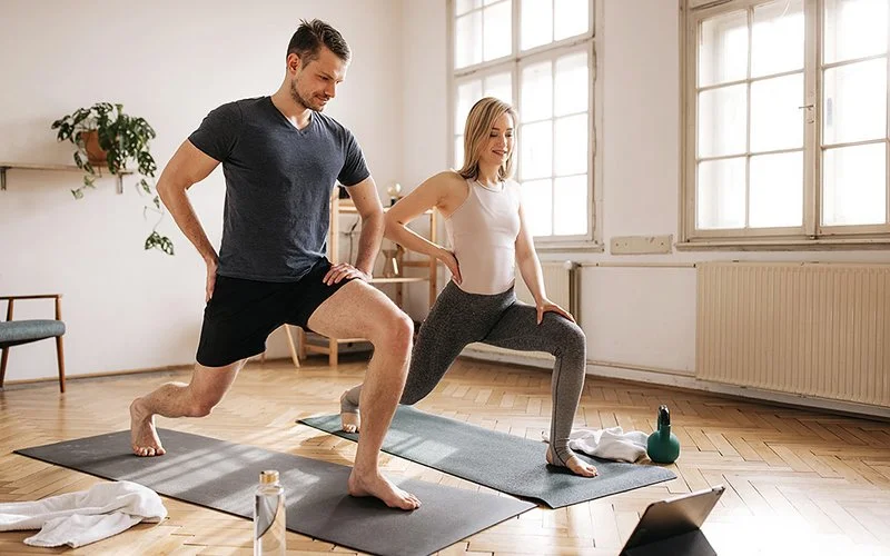 Ein Mann und eine Frau machen zu Hause auf Trainingsmatten Ausfallschritte. Dabei blicken beide auf ein Tablet, das vor ihnen aufgestellt ist.