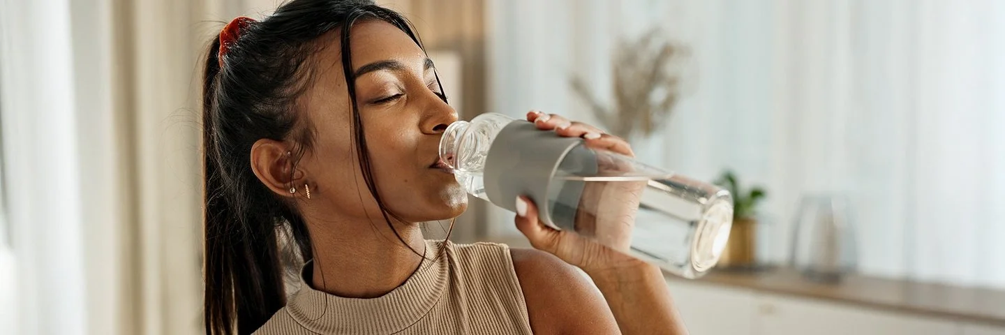 Eine Frau mit einem Zopf und einem brauen Oberteil trinkt Wasser aus einer Glasflasche.