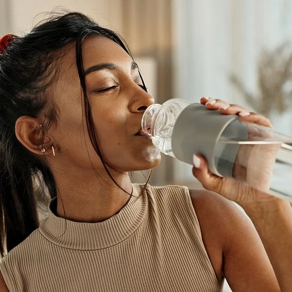 Eine Frau mit einem Zopf und einem brauen Oberteil trinkt Wasser aus einer Glasflasche.