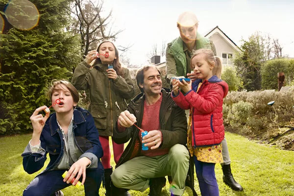 Zu sehen ist eine Familie mit Mutter, Vater und drei Kindern. Sie befinden sich in einem Garten und pusten Seifenblasen in die Luft.