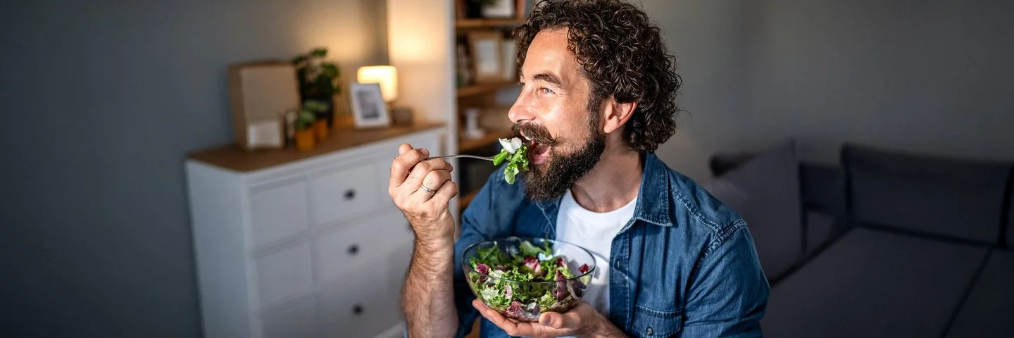 Ein Mann mit Vollbart, offenem Jeanshemd und weißem T-Shirt isst gut gelaunt einen Salat aus einer Glasschüssel.