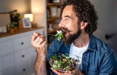Ein Mann mit Vollbart, offenem Jeanshemd und weißem T-Shirt isst gut gelaunt einen Salat aus einer Glasschüssel.