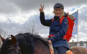 Eine Frau mit blonden Haaren, Helm, blauer Jacke und rotem Rucksack sitzt auf einem Pferd und macht ein Peace-Zeichen.
