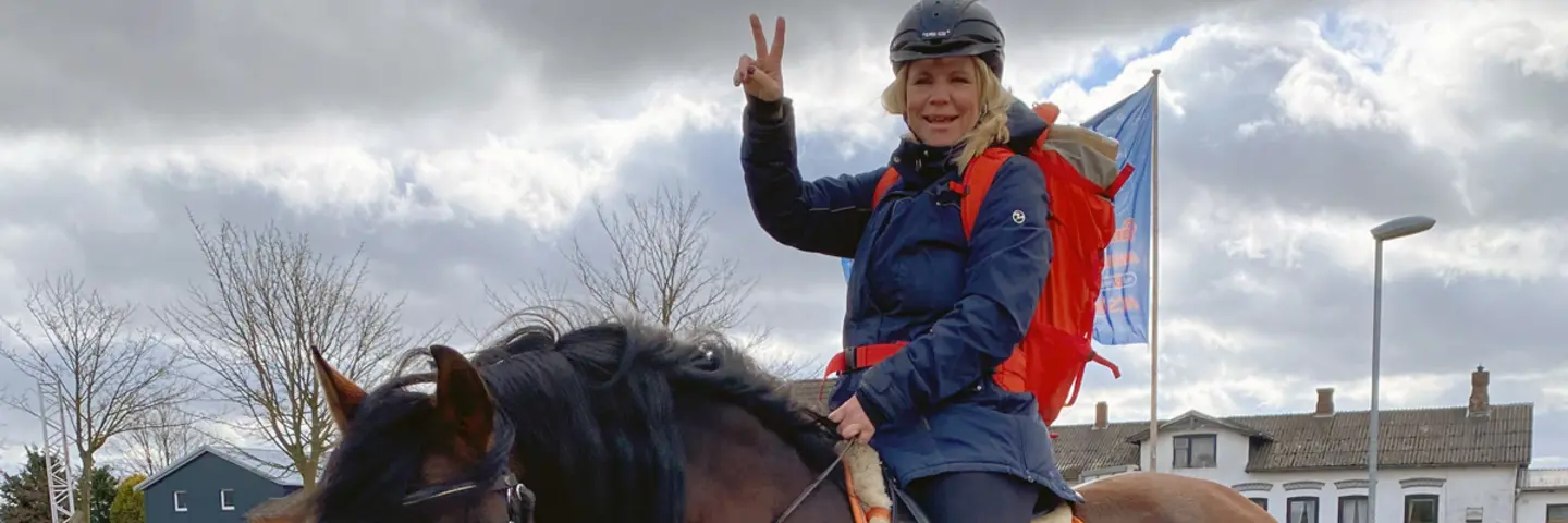 Eine Frau mit blonden Haaren, Helm, blauer Jacke und rotem Rucksack sitzt auf einem Pferd und macht ein Peace-Zeichen.