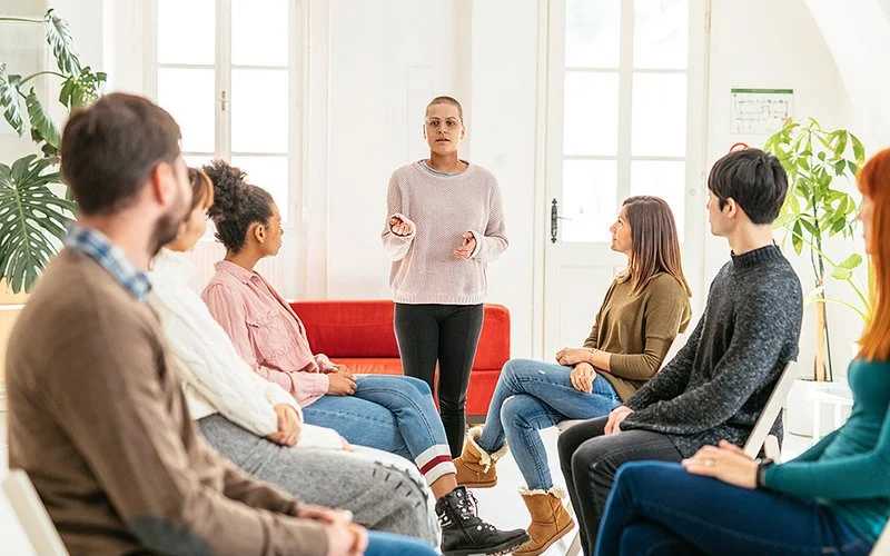 Eine Frau steht vor einer Gruppe von Menschen auf Stühlen, die sie aufmerksam anschauen, die Hände beim Sprechen gehoben.