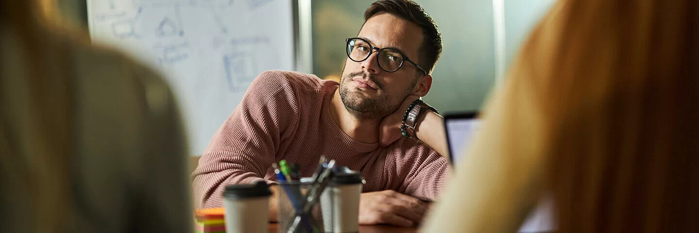 Ein junger Mann sitzt gelangweilt während einer beruflichen Besprechung an einem Bürotisch.