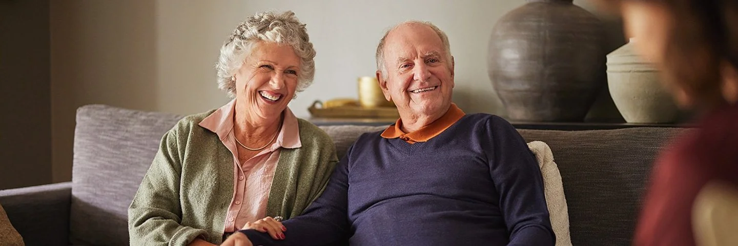 Ein älteres Ehepaar sitzt in einem gemütlichen Wohnzimmer auf einer Couch. Sie lachen und unterhalten sich mit einer Frau, die ihnen gegenüber sitzt. Die Frau notiert etwas auf Papier.