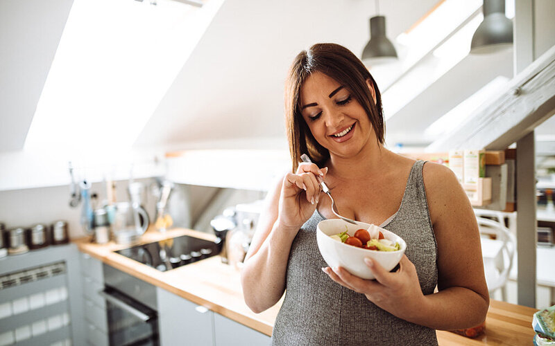 Schwangere Frau isst einen Salat und ernährt sich gesund, um auch in der Schwangerschaft auf ein gesundes Gewicht zu achten.