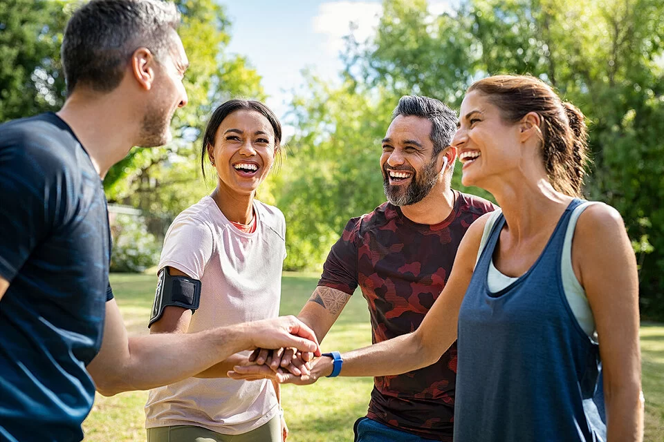 Zwei Männer und zwei Frauen stehen im Park lächelnd beisammen und legen die Hände übereinander. Die AOK-SportTreffs in Baden-Württemberg sind sehr beliebt.