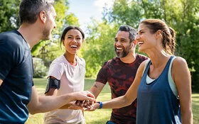 Zwei Männer und zwei Frauen stehen im Park lächelnd beisammen und legen die Hände übereinander. Die AOK-SportTreffs in Baden-Württemberg sind sehr beliebt.