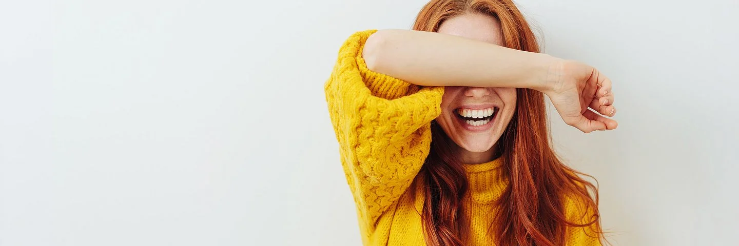 Eine junge Frau im gelben Pulli steht lachend vor einer weißen Wand und legt sich einen Unterarm vor die Augen.