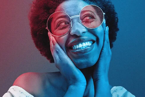 Eine Frau mit Sonnenbrille legt ihr Gesicht in ihre Hände und lächelt in die Kamera.