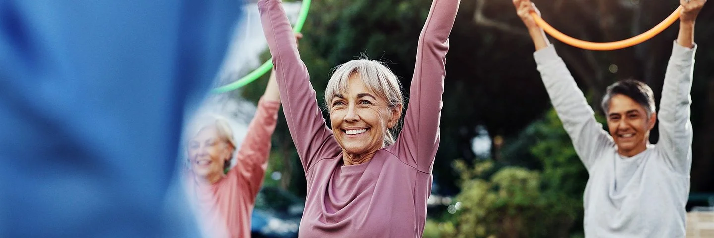 Eine ältere Person mit grauen Haaren trägt ein langärmliges, rosafarbenes Oberteil und hält einen bunten Gymnastikreifen über den Kopf. Im Hintergrund sind weitere Personen mit Gymnastikreifen zu sehen. Die Szene spielt im Freien bei Tageslicht.