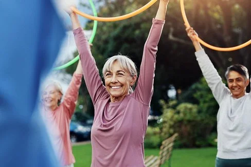 Eine ältere Person mit grauen Haaren trägt ein langärmliges, rosafarbenes Oberteil und hält einen bunten Gymnastikreifen über den Kopf. Im Hintergrund sind weitere Personen mit Gymnastikreifen zu sehen. Die Szene spielt im Freien bei Tageslicht.