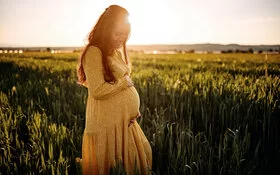 Eine junge, schwangere Frau steht in einem Feld und hält sich lächelnd den Bauch.
