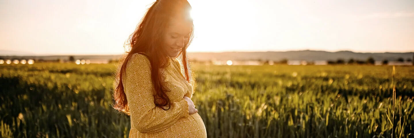 Eine junge, schwangere Frau steht in einem Feld und hält sich lächelnd den Bauch.