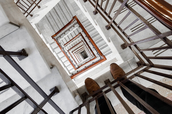 Mann mit Höhenangst steht an einem hohen Treppenhausgeländer und schaut nach unten.