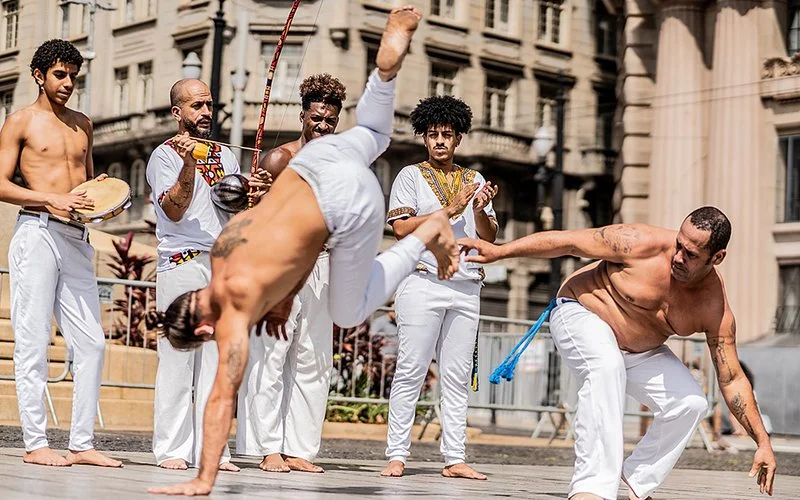Sechs Männer spielen Capoeira auf einem Platz in São Paulo.