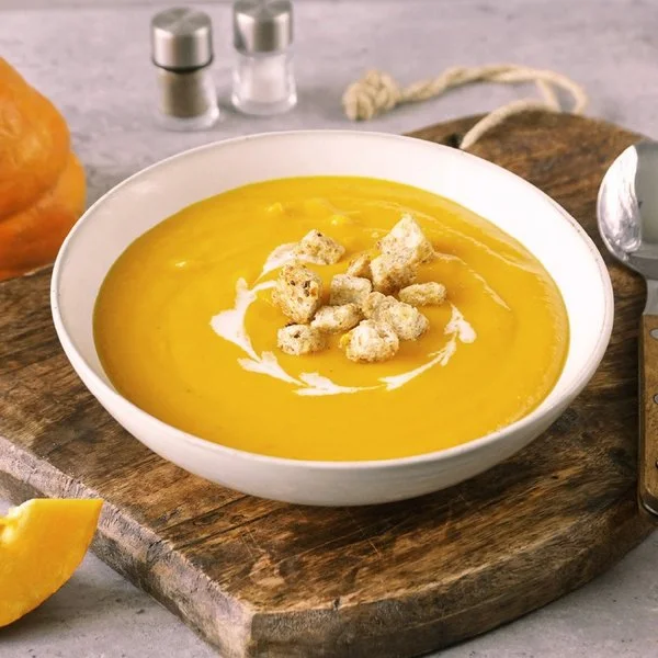 Weißer Teller mit cremiger, oranger Kürbissuppe und Croutons darauf. Der Untergrund besteht aus einem Holzbrett, rechts liegt ein Löffel. Im Hintergrund Möhren, Kürbis und eine weiße Schale mit Croutons.