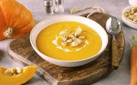 Weißer Teller mit cremiger, oranger Kürbissuppe und Croutons darauf. Der Untergrund besteht aus einem Holzbrett, rechts liegt ein Löffel. Im Hintergrund Möhren, Kürbis und eine weiße Schale mit Croutons.