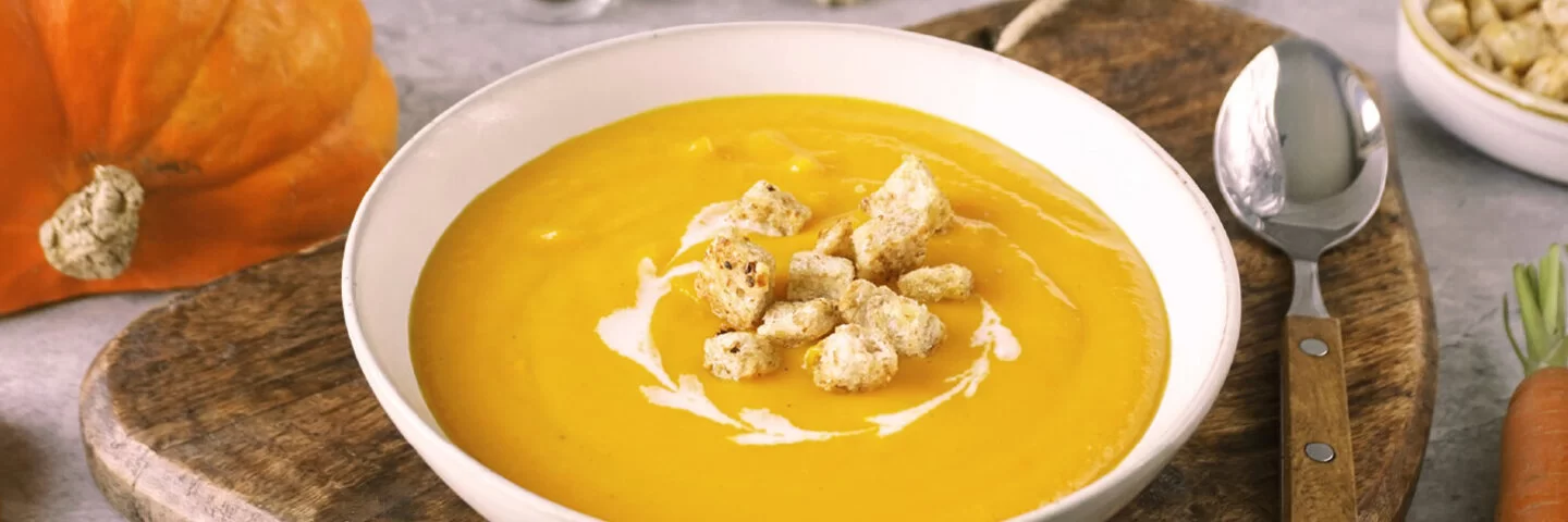 Weißer Teller mit cremiger, oranger Kürbissuppe und Croutons darauf. Der Untergrund besteht aus einem Holzbrett, rechts liegt ein Löffel. Im Hintergrund Möhren, Kürbis und eine weiße Schale mit Croutons.