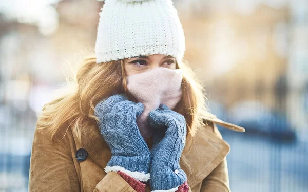 Eine Frau, die sich im Winter draußen mit dicker Winterjacke, Mütze, Handschuhen und Schal vor den kalten Temperaturen schützt.