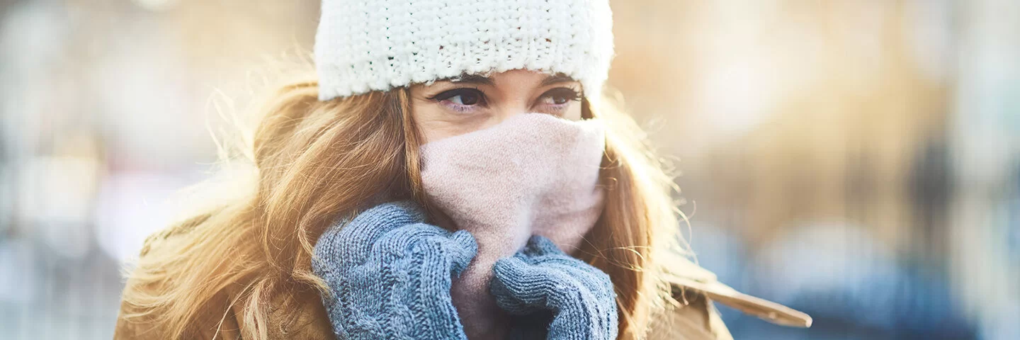 Eine Frau, die sich im Winter draußen mit dicker Winterjacke, Mütze, Handschuhen und Schal vor den kalten Temperaturen schützt.