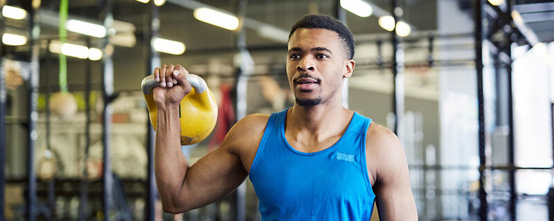 Ein sportlicher Mann Anfang 30 steht in einem Fitnessstudio und hält eine Kettlebell auf Schulterhöhe in seiner rechten Hand.
