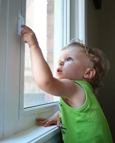 Ein kleiner Junge greift in einer Wohnung an den Griff eines geschlossenen Fensters.