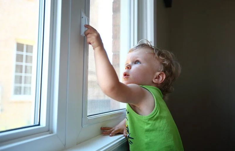 Ein kleiner Junge greift in einer Wohnung an den Griff eines geschlossenen Fensters.