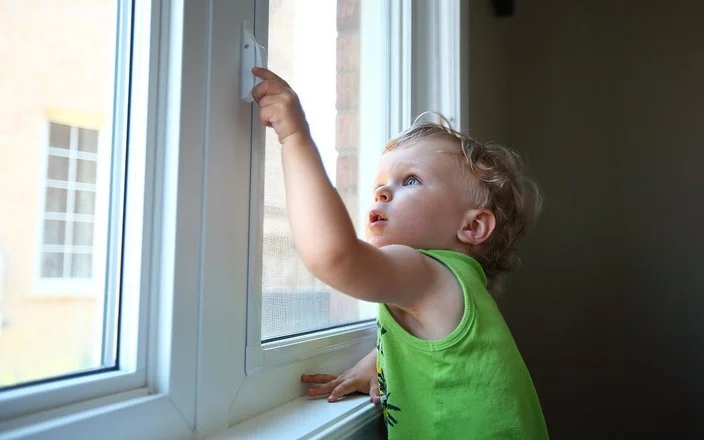 Ein kleiner Junge greift in einer Wohnung an den Griff eines geschlossenen Fensters.
