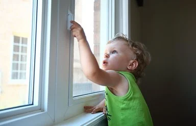 Ein kleiner Junge greift in einer Wohnung an den Griff eines geschlossenen Fensters.