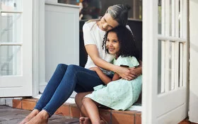 Eine Frau mittleren Alters sitzt mit ihrer ca. sieben Jahre alten Pflegetochter zu Hause in der Terrassentür und umarmt sie.