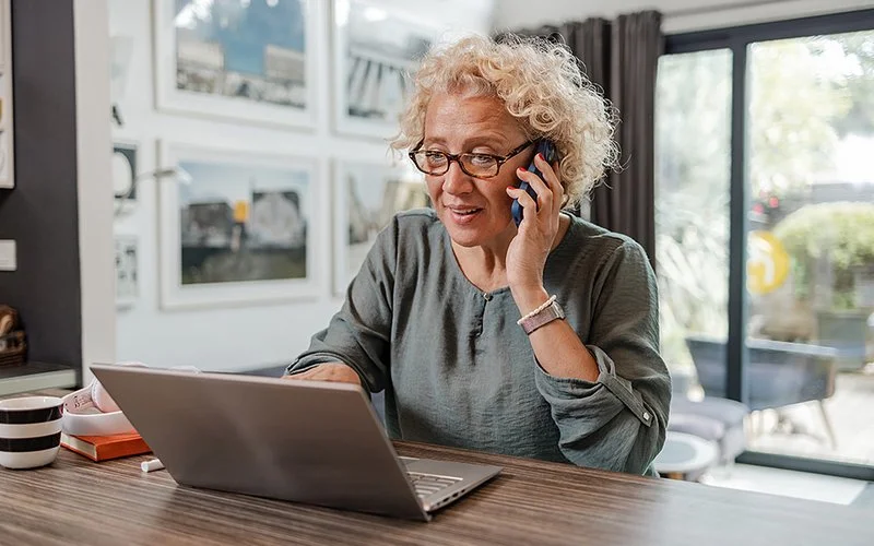 Eine ältere Frau sieht auf den Bildschirm am Laptop und hält ein Telefon in der Hand.