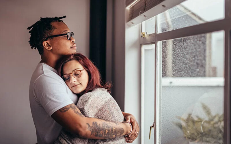 Zwei asexuelle Personen stehen kuschelnd zusammen vor einem Fenster.