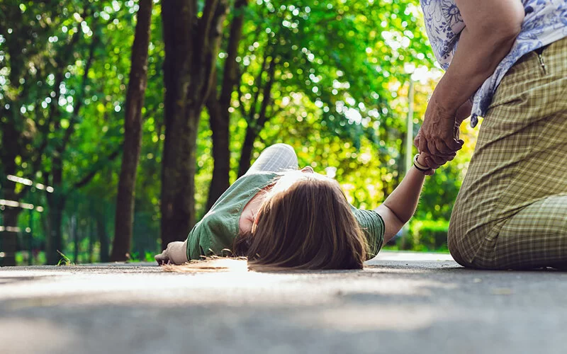 Eine junge Frau liegt ohnmächtig auf einer Straße, eine ältere Frau kniet neben ihr und hält ihre Hand.