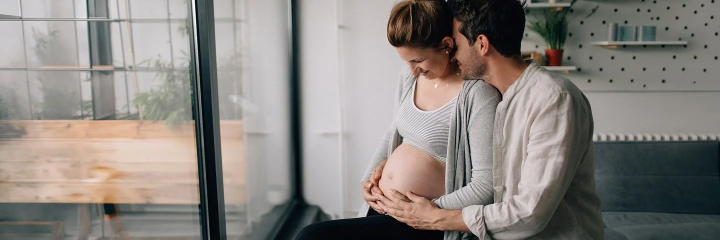 Eine schwangere Frau sitzt vor einem Fenster auf einem Sofa und hält ihren Bauch. Ein Mann sitzt dicht daneben und legt eine Hand auf den Bauch der Schwangeren. 