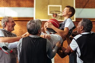 Vier Männer heben einen Jungen hoch, der einen Basketball hält. Hinter ihm ist ein Basketballkorb zu sehen. Alle lachen.