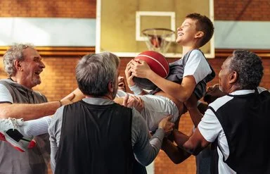 Vier Männer heben einen Jungen hoch, der einen Basketball hält. Hinter ihm ist ein Basketballkorb zu sehen. Alle lachen. 
