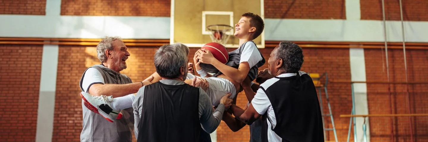 Vier Männer heben einen Jungen hoch, der einen Basketball hält. Hinter ihm ist ein Basketballkorb zu sehen. Alle lachen. 