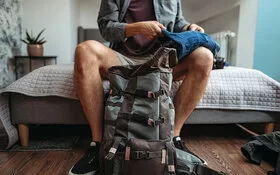 Ein Mann sitzt auf dem Bett und faltet eine Jeanshose. Vor ihm steht ein Rucksack auf dem Boden.