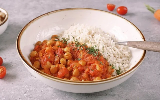 Weißer Teller mit braunem Rand, gefüllt mit rotem Kichererbsencurry mit Reis, bestreut mit Petersilie. Ein Löffel steckt in dem Curry. Im Hintergrund eine Schälchen mit Reis, ein Schälchen mit Kichererbsen, sowie Petersilie, Möhren und Cherrytomaten.