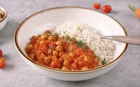 Weißer Teller mit braunem Rand, gefüllt mit rotem Kichererbsencurry mit Reis, bestreut mit Petersilie. Ein Löffel steckt in dem Curry. Im Hintergrund eine Schälchen mit Reis, ein Schälchen mit Kichererbsen, sowie Petersilie, Möhren und Cherrytomaten.