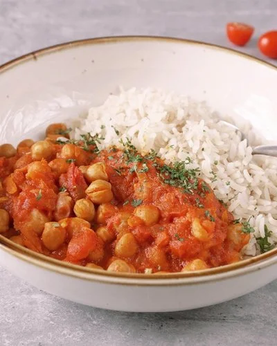 Weißer Teller mit braunem Rand, gefüllt mit rotem Kichererbsencurry mit Reis, bestreut mit Petersilie. Ein Löffel steckt in dem Curry. Im Hintergrund eine Schälchen mit Reis, ein Schälchen mit Kichererbsen, sowie Petersilie, Möhren und Cherrytomaten.