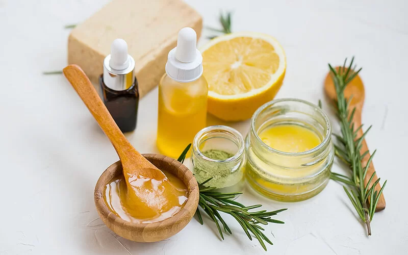 Natürliche Hautpflegezutaten auf einem Tisch: Manuka-Honig, frische Zitrone, ätherisches Öl, Tonerde, pflegender Balsam, Rosmarinzweige und handgemachte Naturseife.
