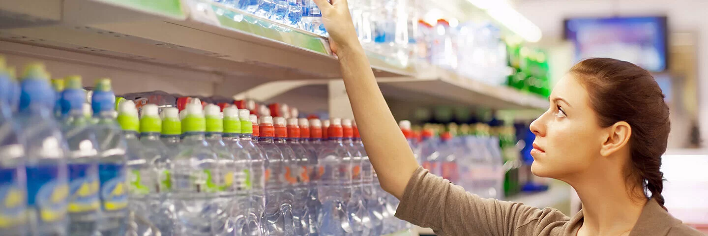 Eine Frau steht vor einem Getränkeregal mit Wasserflaschen in verschiedenen Verpackungen.